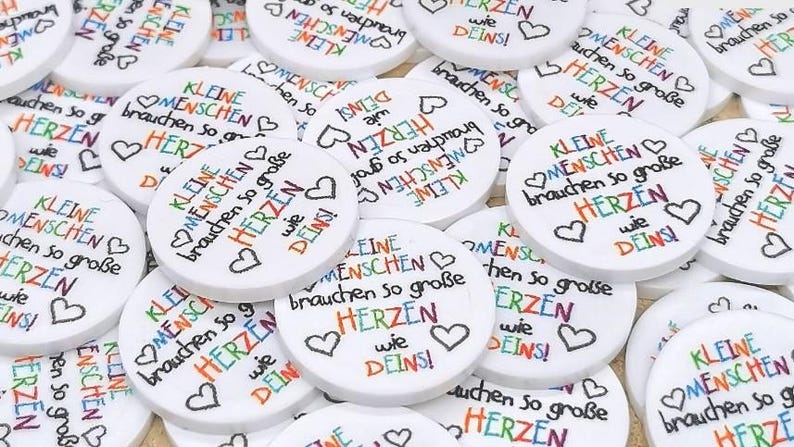 Puede incluir: Una colecci&oacute;n de objetos circulares blancos con la frase alemana "Kleine Menschen brauchen so gro&szlig;e Herzen wie Deins!" en varios colores. Cada disco tiene un s&iacute;mbolo de coraz&oacute;n.