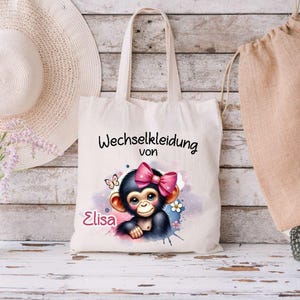 Könnte beinhalten: Eine weiße Stofftasche mit einer rosa Schleife und einem Comic-Affen mit dem Text "Wechselkleidung von Elisa" bedruckt.