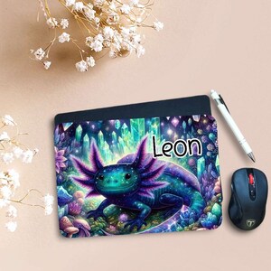 Könnte beinhalten: Ein personalisiertes Mauspad mit einer farbenfrohen Illustration eines Axolotl in Blau-, Lila- und Grüntönen, mit dem Namen "Leon". Eine schwarze Computermaus und ein Stift liegen neben dem Mauspad. Der Hintergrund zeigt Kristalle.