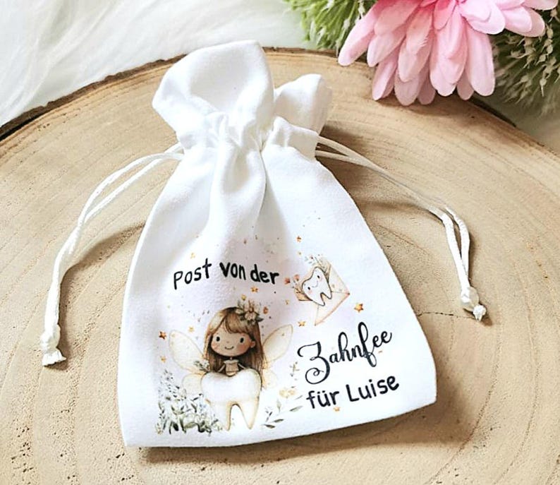 May include: A white fabric bag with a drawstring closure. The bag has a printed design of a tooth fairy with a tooth, flowers, and stars. The text on the bag reads "Post von der Zahnfee f&uuml;r Luise".