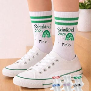 Könnte beinhalten: Weiße Socken mit grünen Streifen, dem Text "Schulkind 2026 Nelio" und einer Regenbogen-Grafik. Getragen mit weißen Leinenschuhen. Zusätzliche Socken mit farbigen Streifen werden angezeigt.