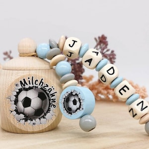Könnte beinhalten: Eine helle Holzdose für Milchzähne mit Deckel, verziert mit einem Fußballmotiv und dem Schriftzug "Milchzähne". Daran befestigt ist eine Kette aus hellblauen, grauen und naturfarbenen Holzperlen mit Würfeln, die den Namen "JADEN" bilden.