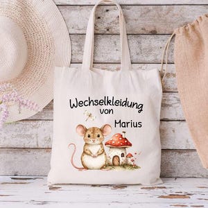 Könnte beinhalten: Eine weiße Stofftasche mit dem Text "Wechselkleidung von Marius" und einer Comic-Illustration einer Maus und einem Pilzhaus.
