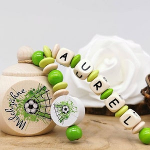 Könnte beinhalten: Holz-Schnullerband mit dem Namen "AUREL" in schwarzen Buchstaben auf Holzblöcken. Der Clip hat grüne und hellgrüne Perlen und ein Fussball-Design. Eine kleine Holzbox ist ebenfalls im Bild.