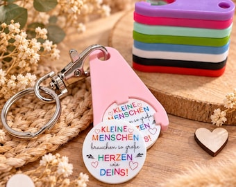Könnte beinhalten: Ein rosa Kunststoff-Schlüsselanhänger mit einem runden weißen Einsatz, der den Text KLEINE MENSCHEN brauchen so große HERZEN wie DEINS! in bunten Buchstaben zeigt, begleitet von einem Stapel aus sechs weiteren bunten Kunststoff-Haltern.