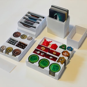 May include: White plastic organizer with compartments for storing game pieces and cards. The organizer has compartments for coins, tokens, and cards. The organizer is designed to store game components for a board game.