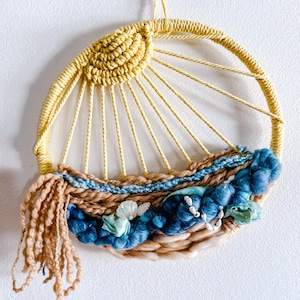 May include: A handcrafted wall hanging featuring a yellow sun design with radiating lines. The lower section incorporates blue and tan woven textures, accented with light blue fabric flowers and white beads. The circular piece is suspended by a thin yellow cord.
