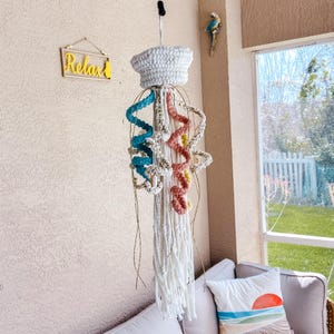 May include: A handmade, white macrame jellyfish mobile with blue, pink, and yellow accents. The jellyfish has a crocheted top and long, flowing tentacles. The word "Relax" is on a small sign in the background.