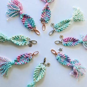 May include: A collection of handmade macrame keychains in a circular arrangement. Each keychain features a braided design in pastel colors of pink, blue, and green, with a tassel at the end. Some have charms like stars or compasses.