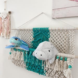 May include: A macrame wall hanging storage organizer featuring cream and teal cotton cord knots, suspended from a wooden dowel. The organizer holds a blue plush sea turtle and a grey plush manatee, with decorative fringe along the bottom edge.