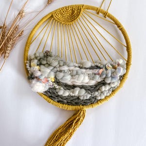 May include: A round, woven wall hanging with a yellow sunburst design. The center features textured gray and white yarn with pink and iridescent accents. A yellow tassel hangs from the bottom, and the piece is approximately 10 inches in diameter.