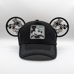 May include: Black trucker hat featuring a Mickey Mouse patch and mouse ear accents. The hat has a mesh back and a curved brim. The Mickey Mouse patch is square and has the word "Mickey" above the image. The mouse ear accents feature Mickey Mouse holding a ship's wheel.