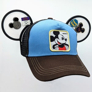 May include: A blue and brown trucker hat with a Mickey Mouse patch that says "Mickey" on it. The hat has a mesh back and a brown brim.