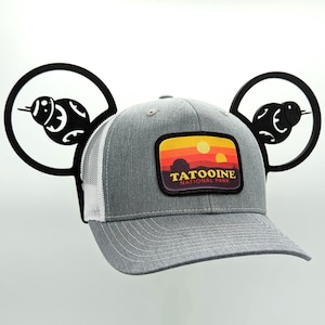 Może przedstawiać: Szara i biała czapka trucker z czarną naszywką z napisem "Tatooine National Park" i sceną zachodu słońca z dwoma słońcami. Czapka ma czarne uszy Myszki Miki z sylwetką Gwiazdy Śmierci na każdym uchu.