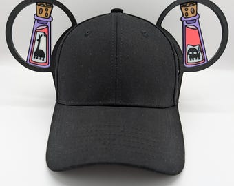 May include: Black baseball cap with two black ears shaped like bottles with purple and pink labels. The labels have a skull and crossbones and a llama on them.