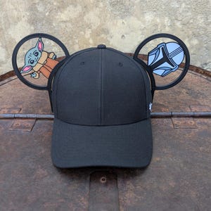 May include: Black baseball cap with a black and white Mandalorian helmet and a green Baby Yoda character on the sides.