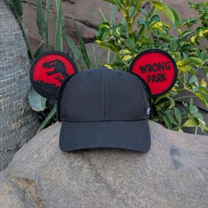 May include: A black baseball cap with red and black Mickey Mouse ears. The left ear has a silhouette of a dinosaur with the text "WRONG PARK" on the right ear.