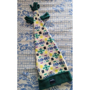 May include: A cream-colored fleece hat with a geometric pattern in green, blue, yellow, and purple. The hat has a green brim and green tassels at the top. The hat is approximately 12 inches long.