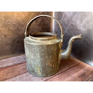 May include: A vintage copper kettle with a patina finish and a handle. The kettle has a spout and is sitting on a wooden surface.
