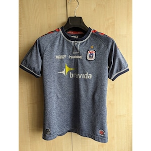 Può includere: Una maglia da calcio blu e grigia con accenti rossi e bianchi. La maglia ha i loghi di Hummel, Window Master e una squadra chiamata "MIF". Il testo "bravida" è stampato sul davanti della maglia.