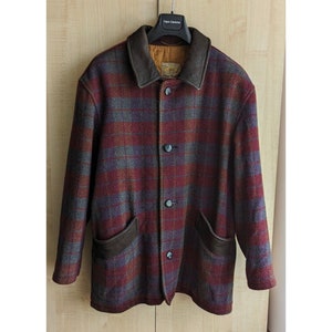 Puede incluir: Una chaqueta de cuadros rojos, grises y azules con cuello y bolsillos de cuero marrón. La chaqueta tiene cierre de botones y está hecha de una tela gruesa y cálida.