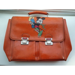 May include: Orange vintage school bag featuring a Mickey Mouse graphic. The bag has a handle, two metal clasps, and a stitched design around the edges. The bag is rectangular and has two front pockets.