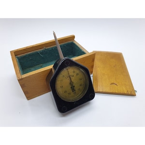 Könnte beinhalten: Ein schwarz-weißes Messgerät mit einer Holzkiste. Das Messgerät hat ein schwarzes Zifferblatt mit weißen Markierungen und den Text "ГРОМ" in kyrillischer Schrift. Die Kiste ist aus hellem Holz und hat eine grüne Filz-Auskleidung.