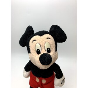 May include: A plush Mickey Mouse toy with black ears, a black nose, and large, round eyes. The toy is wearing red shorts with a black button.