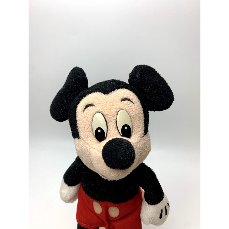 May include: A plush Mickey Mouse toy with black ears, a black nose, and large, round eyes. The toy is wearing red shorts with a black button.