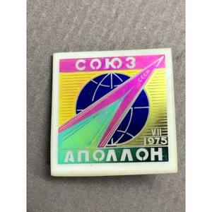 May include: A square pin featuring Cyrillic text "СОЮЗ" and "АПОЛЛОН", a blue globe, and a pink and green arrow design. The pin also includes the text "CCCP" and "USA" and the date "VII 1975".