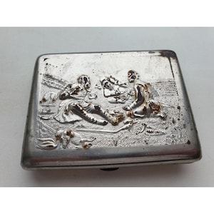 May include: A silver metal cigarette case with an embossed design of three people sitting on the ground. The design is detailed and includes a dog and a gun.