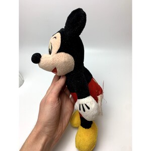 May include: A vintage plush Mickey Mouse toy with black fur, red shorts, and yellow shoes. The toy has a worn and faded appearance, suggesting it is an older item.