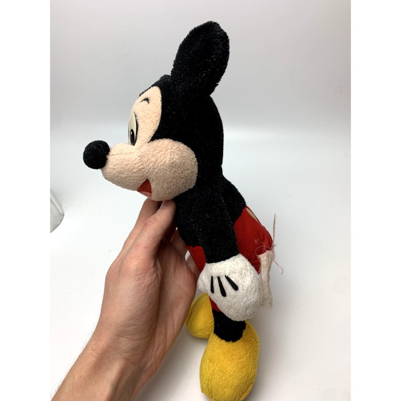 May include: A vintage plush Mickey Mouse toy with black fur, red shorts, and yellow shoes. The toy has a worn and faded appearance, suggesting it is an older item.