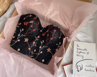 May include: A black corset with floral embroidery and butterfly details. The corset is in a gift box with tissue paper and a book titled "The Unbearable Lightness of Being" by Milan Kundera.