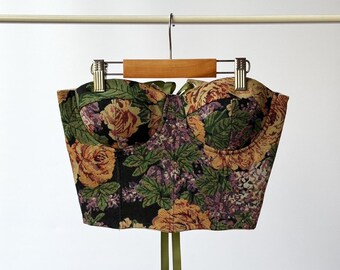 May include: A black floral print corset with green satin ribbon ties. The corset has a sweetheart neckline and is made of a tapestry fabric.