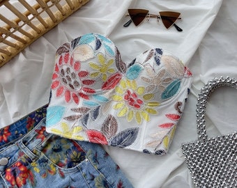 May include: A white floral embroidered bustier top with a sweetheart neckline. The top has a colorful floral pattern with red, yellow, blue, and green accents.