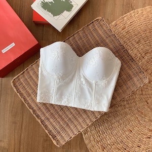 Op de afbeelding: Witte strapless corset met delicate bloemenborduursels. Het corset heeft een beenste structure en een gladde, satijnachtige afwerking.