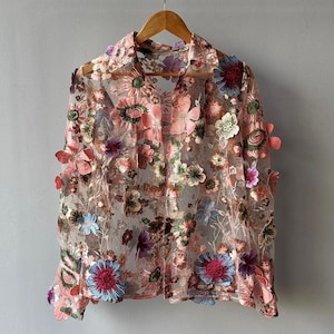 May include: A sheer, long-sleeved blouse with a floral embroidered design. The blouse is pink with multi-coloured flowers.