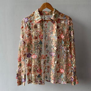 Op de afbeelding: Een doorschijnende, langarmige blouse met een bloemenborduurpatroon in tinten roze, rood, paars en groen. De blouse heeft een kraag en een knoopsluiting.