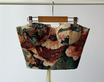 May include: A floral tapestry corset top with a sweetheart neckline. The top features a mix of colors including burgundy, orange, green, and cream. It hangs from a wooden hanger with metal clips, and has cream-colored ribbon ties.