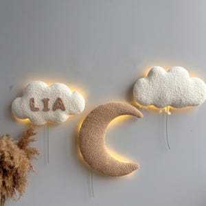 Pode incluir: Três luzes decorativas de parede em forma de nuvem, lua e outra nuvem. As nuvens são brancas e a lua é castanha. A nuvem da esquerda tem o nome "LIA". As três luzes têm um brilho quente.