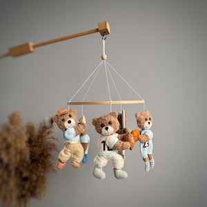 Può includere: Un mobile per bambini in legno con quattro orsacchiotti marroni che indossano diverse uniformi sportive. Gli orsacchiotti tengono una mazza da baseball, un pallone da calcio, una palla da basket e una mazza da golf. Il mobile è appeso a un braccio di legno.