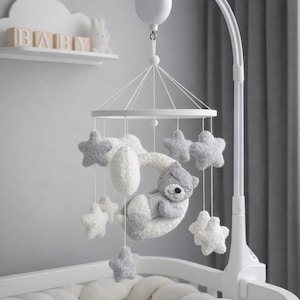 Puede incluir: Una cuna blanca con un parachoques trenzado y un móvil colgante. El móvil presenta un oso de peluche gris y blanco, nubes blancas y estrellas grises y blancas. Una estantería sobre la cuna muestra la palabra "BABY".