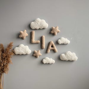 May include: A nursery decor set with the name "LIA" spelled out in beige felt letters, surrounded by white felt clouds and beige felt stars.