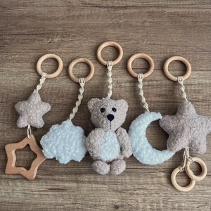 May include: A baby mobile with a wooden star, a plush cloud, a teddy bear, a plush crescent moon, and a plush star. The mobile has wooden rings and is suspended by a cream-coloured string. The items are in neutral colours.