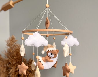 May include: A wooden baby mobile with a brown bear sleeping on a crescent moon, white clouds, and beige stars. The mobile is suspended from a wooden arm.