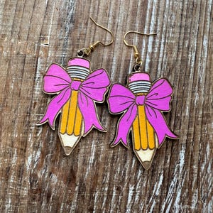 May include: Wooden dangle earrings shaped like pencils with pink bows. The pencils are yellow with a brown tip and a pink eraser. The earrings have gold-colored hooks.