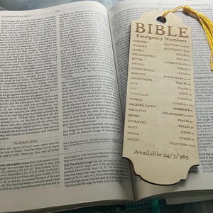 Może przedstawiać: Otwarta Biblia z drewnianą zakładką. Zakładka zawiera „NUMERY ALARMOWE BIBLII” i powiązane wersety biblijne dla różnych stanów emocjonalnych. Żółty chwost zdobi zakładkę. Tekst „Dostępne 24/7/365” jest wydrukowany na dole.
