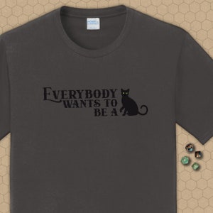 May include: A dark gray t-shirt with the text "Everybody wants to be a" and a black cat with yellow eyes sitting on the right side of the text.