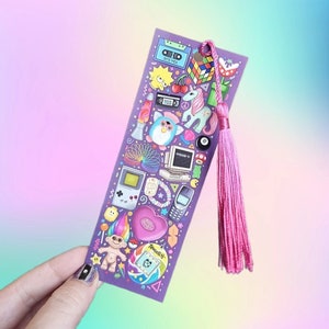 Pode incluir: Um marcador de livros roxo com ilustra&ccedil;&otilde;es dos anos 90, incluindo um Cubo de Rubik, Furby, Game Boy e um Tamagotchi. O marcador de livros tem uma borla rosa. O fundo &eacute; um gradiente de rosa, roxo e azul.
